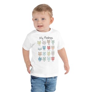 May include: A white toddler t-shirt with a graphic of cats with different facial expressions. The text "My Feelings" is printed above the cats. Each cat has a word underneath it describing a feeling, such as happy, sad, scared, and angry.