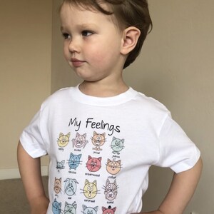 May include: White t-shirt with the text "My Feelings" and a graphic of 18 cartoon cats with different facial expressions representing various emotions. Each cat has a word describing the emotion below it.