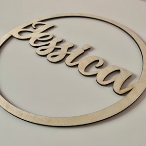 May include: A wooden sign featuring the name "Jessica" in a cursive font, enclosed within a circular frame. The sign is crafted from light-coloured wood with a dark edge, contrasting against the white background. The design is simple and elegant.