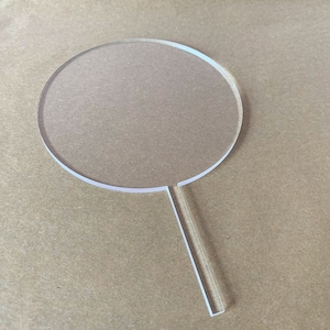 May include: A clear acrylic cake topper in the shape of a circle with a long, thin handle. The circle is about 4 inches in diameter.