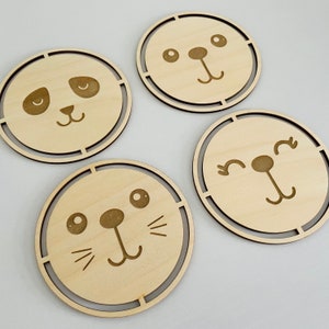 May include: Four wooden coasters with different animal faces. Each coaster features a simple line drawing of a panda, a cat, a dog, and a seal. The coasters are all round and have a natural wood finish.