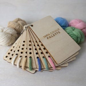 May include: A wooden yarn colour palette with the text "YARN COLOUR PALETTE" engraved on the front. The palette is attached to a metal ring and displays various yarn colours, including white, beige, green, blue, and pink. Several yarn balls are in the background.