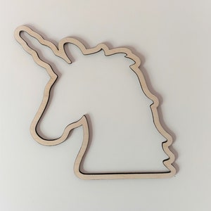 May include: A wooden unicorn head outline, featuring a natural wood grain finish. The unicorn's profile is cut out, showcasing the shape of the horn, mane, and face. The cutout is set against a white background, highlighting the detailed craftsmanship of the wooden art piece.