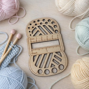 May include: A wooden yarn gauge and ruler with various sized holes and a ruler marked in inches and centimeters. The gauge is surrounded by balls of yarn in pastel colors, with knitting needles and a small knitted swatch.