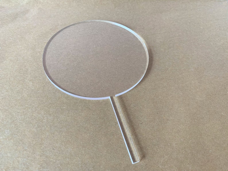 May include: A clear acrylic cake topper in the shape of a circle with a long, thin handle. The circle is about 4 inches in diameter.