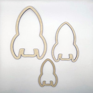 May include: Three wooden rocket ship cutouts in varying sizes. The rockets are light brown with a dark outline, and are arranged on a white background. The largest rocket is approximately 6 inches tall.