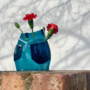 Könnte beinhalten: Eine tealblaue Glasvase mit einem einzigartigen Design, mit einer Tasche auf der Vorderseite. Die Vase ist mit roten Nelken gefüllt.