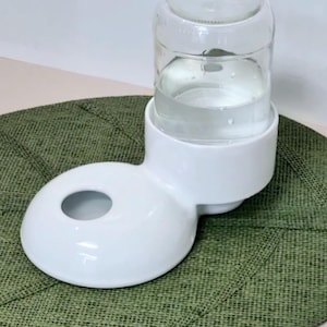 Könnte beinhalten: Ein weißer Keramik-Wasserspender für Haustiere mit einer glatten, abgerundeten Basis mit kreisförmiger Öffnung und einer klaren Glasflasche, die in einen zylindrischen Behälter umgedreht ist. Die Struktur hat eine saubere, glänzende Oberfläche.