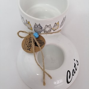 Op de afbeelding: Witte keramische dubbele kom met kattenmotief en een bruin papieren label met de tekst "Daisy Ceramics". De kom heeft een ring met katten rond de bovenkant en de woorden "Cat's" op de bodem.
