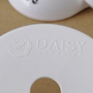Op de afbeelding: Witte keramische basis met een gat in het midden. De basis heeft een reli&euml;flogo met de tekst "Daisy Ceramics" en een gestileerd madeliefje-ontwerp.
