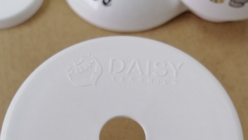 Op de afbeelding: Witte keramische basis met een gat in het midden. De basis heeft een reli&euml;flogo met de tekst "Daisy Ceramics" en een gestileerd madeliefje-ontwerp.
