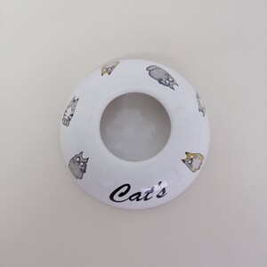 May include: White ceramic ring holder with a cat design and the word "Cat's" printed on the bottom.