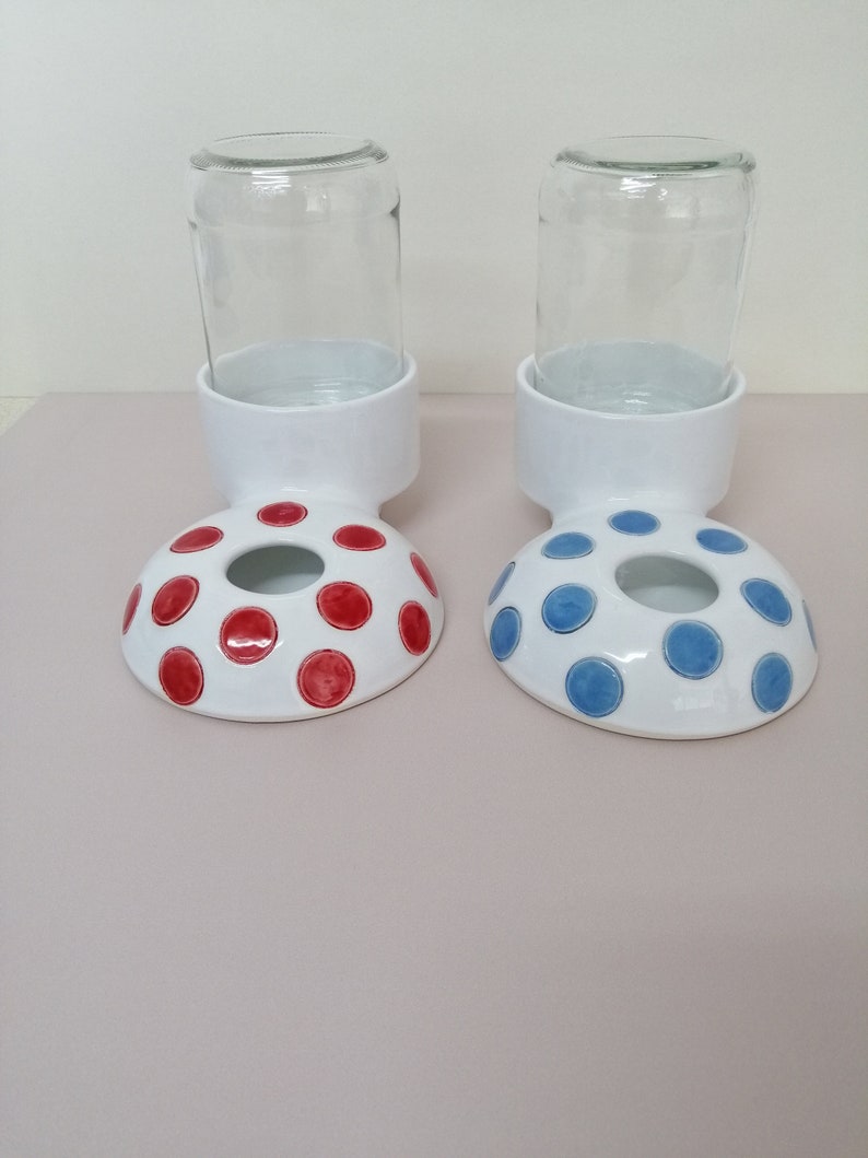 May include: Two white ceramic pet food and water bowls with red and blue polka dots. Each bowl has a clear glass jar on top.