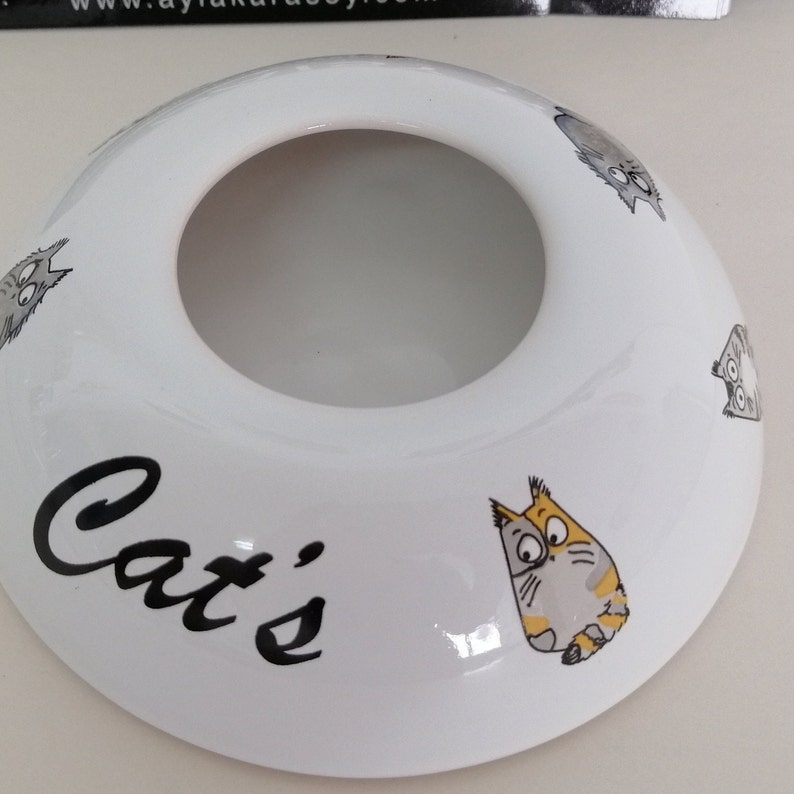 May include: White ceramic cat-themed dish with a hole in the center. The dish has the word "Cat's" written in black cursive. There are several cartoon cats drawn on the dish.