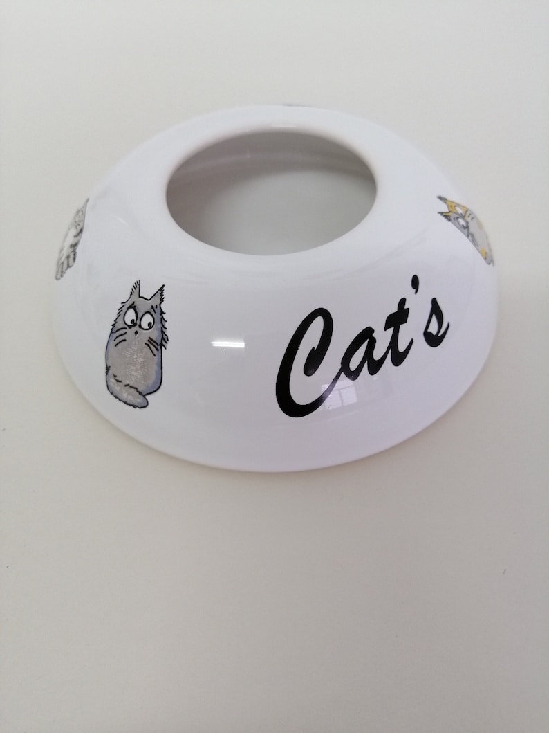 May include: White ceramic cat dish with a hole in the center and the word "Cat's" written in black. The dish is decorated with cartoon cats.