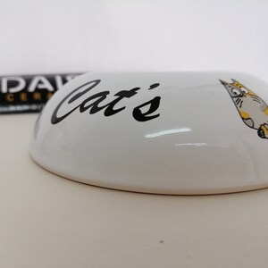 May include: White ceramic cat food bowl with black lettering that says "Cat's" and a cartoon cat illustration.