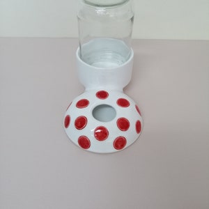 May include: White ceramic pet water dispenser with red polka dots. The dispenser has a hole in the top for a glass jar to be placed inside.