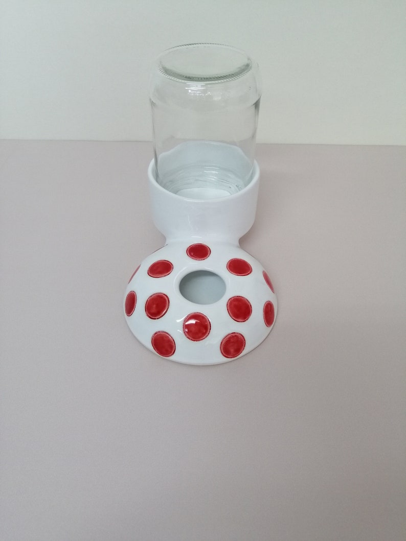 May include: White ceramic pet water dispenser with red polka dots. The dispenser has a hole in the top for a glass jar to be placed inside.