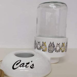 Op de afbeelding: Witte keramische kattenvoedsel dispenser met een glazen pot erbovenop. De dispenser heeft een kom met het woord "Cat's" erop. De pot heeft een rij cartoon katten rond de onderkant.