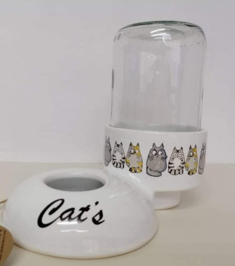 Op de afbeelding: Witte keramische kattenvoedsel dispenser met een glazen pot erbovenop. De dispenser heeft een kom met het woord "Cat's" erop. De pot heeft een rij cartoon katten rond de onderkant.