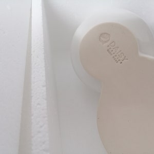 May include: A white ceramic bowl with a heart-shaped design. The bowl has a raised inscription that reads "DAISY CERAMICS HAND MADE"