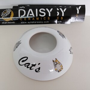 May include: White ceramic cat food bowl with a black inscription that reads "Cat's" and a cartoon cat design. The bowl is on a white surface with a black and yellow sticker that reads "DAISY CERAMICS www.aylakarasoy.com" in the background.
