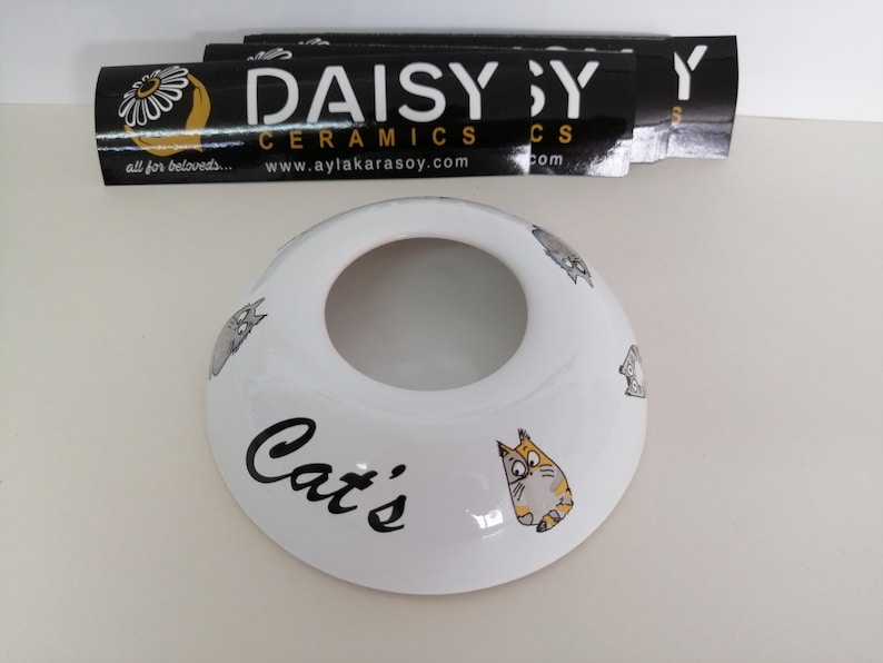 May include: White ceramic cat food bowl with a black inscription that reads "Cat's" and a cartoon cat design. The bowl is on a white surface with a black and yellow sticker that reads "DAISY CERAMICS www.aylakarasoy.com" in the background.