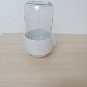 May include: White ceramic pet bowl with a clear glass water bottle holder. The bowl is heart-shaped and has a raised rim around the edge.