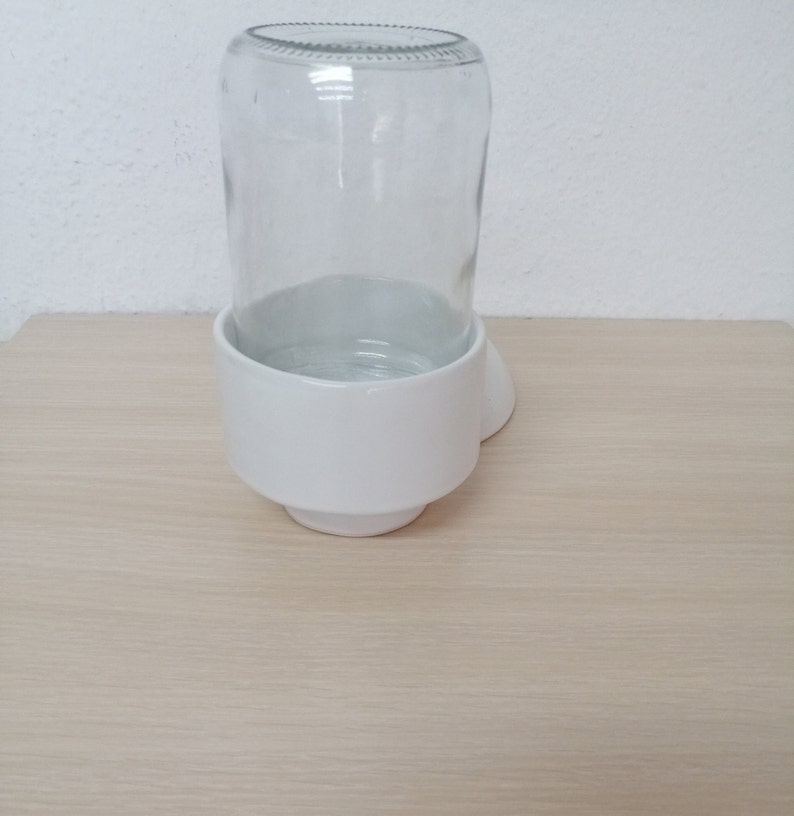 May include: White ceramic pet bowl with a clear glass water bottle holder. The bowl is heart-shaped and has a raised rim around the edge.