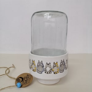 Op de afbeelding: Witte keramische plantenpot met een rij cartoonkatten rond de bovenkant. De pot heeft een transparante glazen cilinder bovenop. De pot heeft een bruin label met de tekst "At The Daisy Ceramics".