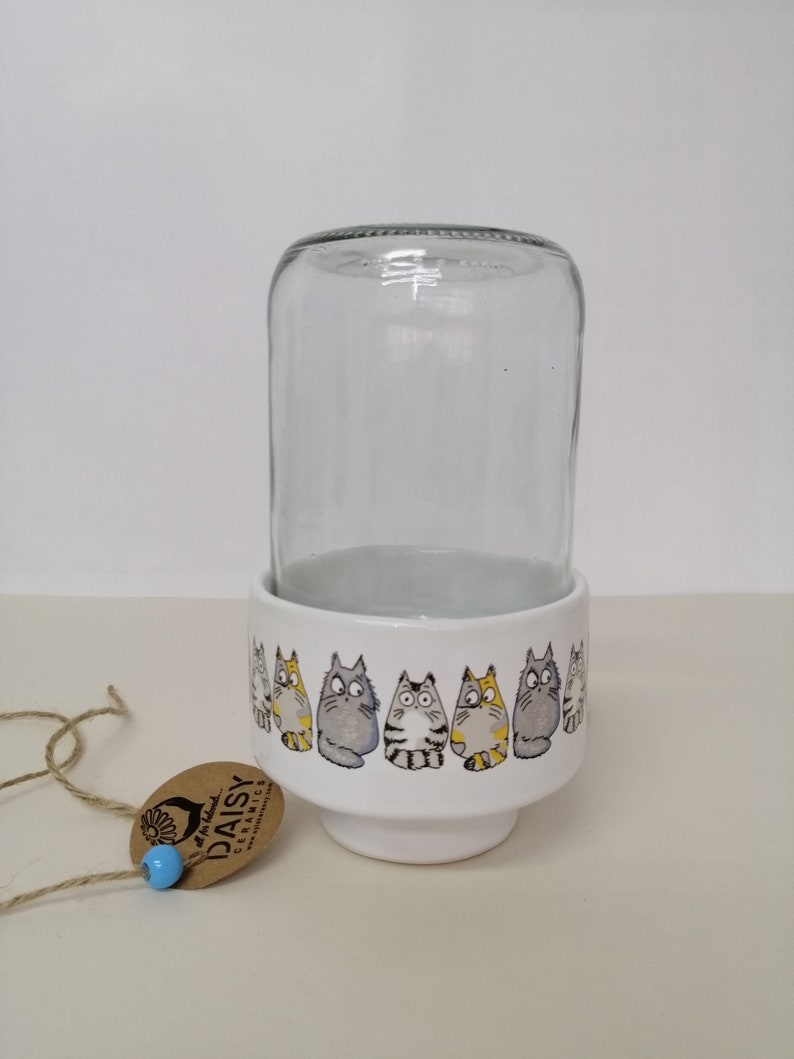 Op de afbeelding: Witte keramische plantenpot met een rij cartoonkatten rond de bovenkant. De pot heeft een transparante glazen cilinder bovenop. De pot heeft een bruin label met de tekst "At The Daisy Ceramics".