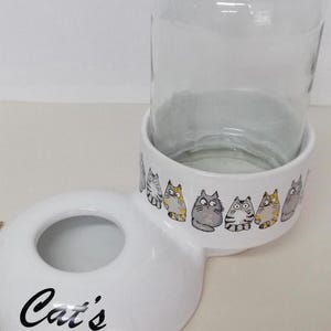 Op de afbeelding: Witte keramische kattenvoer- en waterbakset met een glazen pot voor water. De bak heeft een verhoogde rand en de tekst "Cat's" gedrukt op de voorkant. De pot heeft een witte keramische basis met een rij cartoonkatten gedrukt rond de rand.