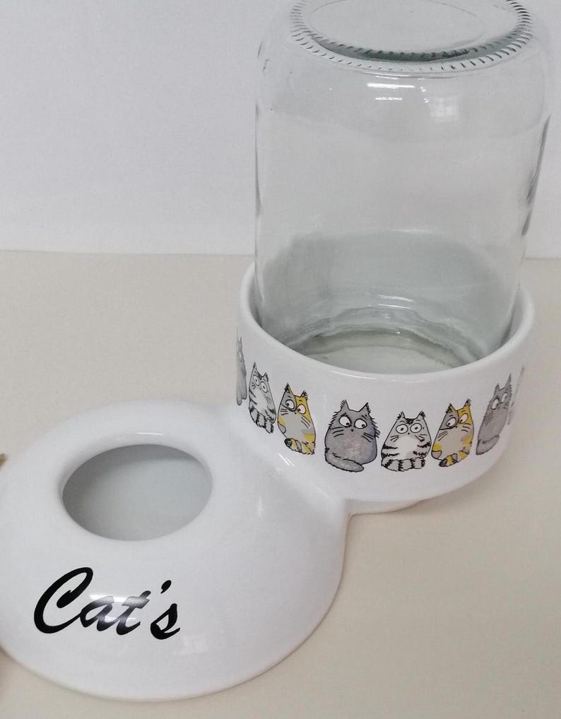 Op de afbeelding: Witte keramische kattenvoer- en waterbakset met een glazen pot voor water. De bak heeft een verhoogde rand en de tekst "Cat's" gedrukt op de voorkant. De pot heeft een witte keramische basis met een rij cartoonkatten gedrukt rond de rand.