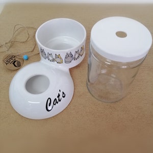 Op de afbeelding: Witte keramische kattenvoer- en waterbakset met een kattenontwerp. De voerbak heeft een aparte waterbak die eraan is bevestigd. De waterbak bestaat uit een glazen pot met een witte deksel.