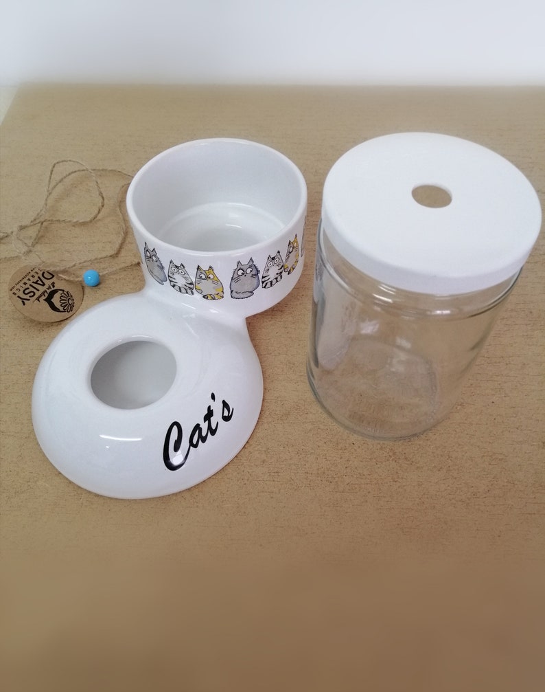 Op de afbeelding: Witte keramische kattenvoer- en waterbakset met een kattenontwerp. De voerbak heeft een aparte waterbak die eraan is bevestigd. De waterbak bestaat uit een glazen pot met een witte deksel.