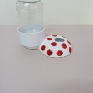 May include: White ceramic pet water dispenser with a red polka dot design. The dispenser has a raised rim to hold a glass jar.
