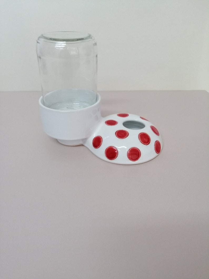 May include: White ceramic pet water dispenser with a red polka dot design. The dispenser has a raised rim to hold a glass jar.