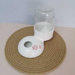 Könnte beinhalten: Ein weißer Keramik-Tränke für Haustiere mit einer durchsichtigen Glasflasche. Der Wasserspender hat eine kreisförmige Basis mit rosa Pfotenabdrücken. Er steht auf einer runden, gewebten Matte.