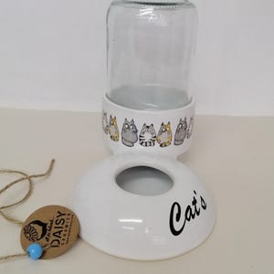 Op de afbeelding: Witte keramische kattenvoedingsdispenser met een glazen pot en een voet die "Cat's" zegt. De voet is versierd met een rij cartoonkatten.