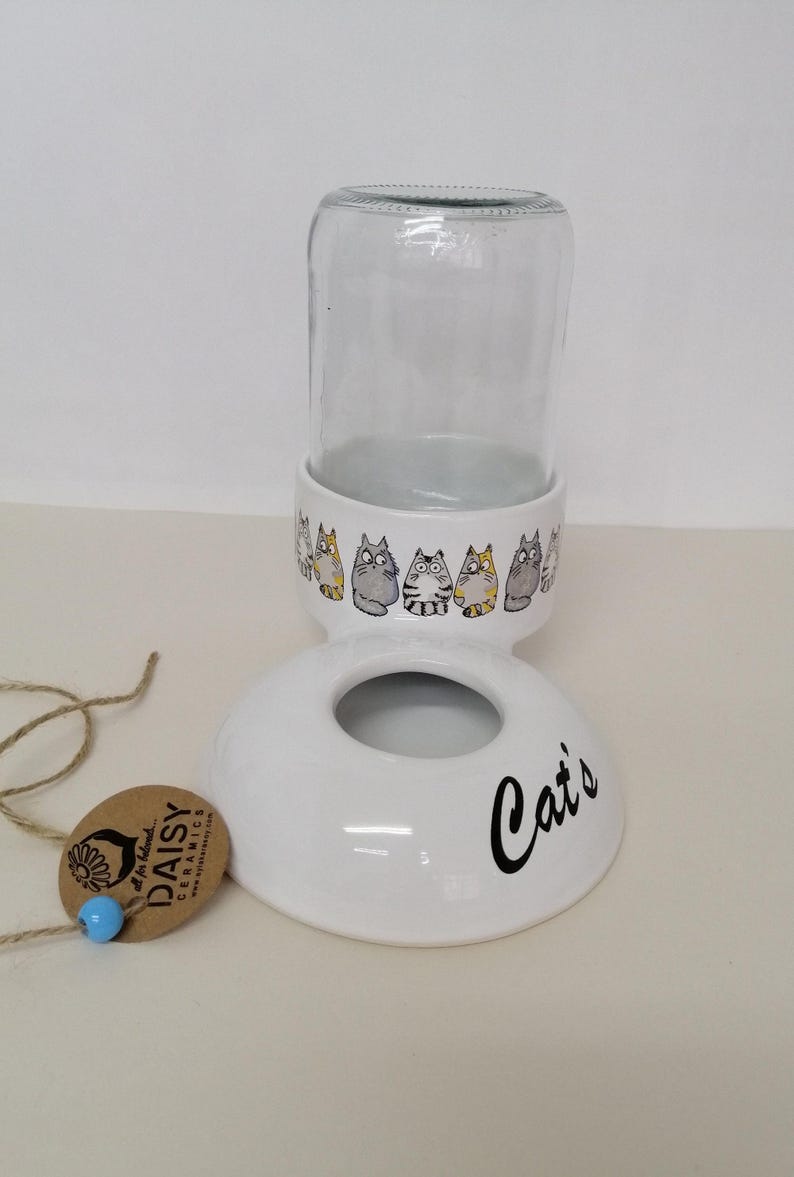 Op de afbeelding: Witte keramische kattenvoedingsdispenser met een glazen pot en een voet die "Cat's" zegt. De voet is versierd met een rij cartoonkatten.