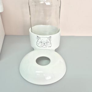 Op de afbeelding: Een drinkfontein voor huisdieren met een doorzichtige glazen fles die in een witte keramische voet is geplaatst. De voet is versierd met een grijze lijntekening van een kattengezicht en bevat een losse witte keramische kom met opening.