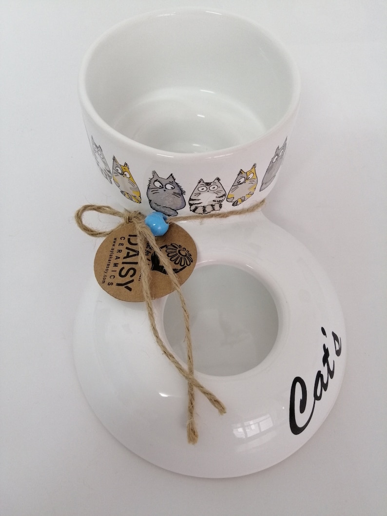 Op de afbeelding: Witte keramische dubbele kom met kattenmotief en een bruin papieren label met de tekst "Daisy Ceramics". De kom heeft een ring met katten rond de bovenkant en de woorden "Cat's" op de bodem.