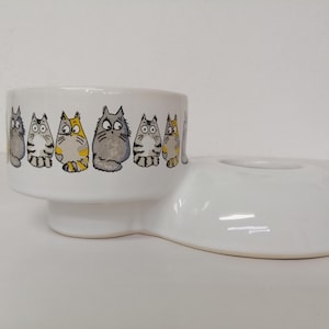 Op de afbeelding: Witte keramische kattenvoederbak met een verhoogde rand en een aparte waterbak. De bak is versierd met een rij cartoonkatten in verschillende poses.