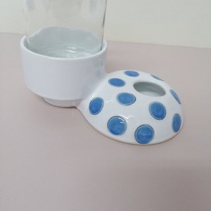 May include: White ceramic bottle holder with a blue polka dot design. The holder has a round base and a raised lip to hold a bottle.