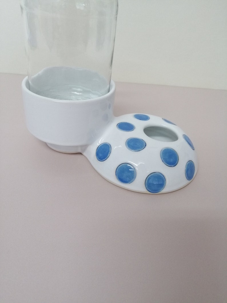 May include: White ceramic bottle holder with a blue polka dot design. The holder has a round base and a raised lip to hold a bottle.