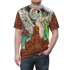 Puede incluir: Una camiseta de manga corta con una ilustración surrealista. El diseño incluye una criatura con forma de ave con alas, ojos grandes y un paisaje urbano. La paleta de colores está dominada por tonos de marrón, naranja, verde y blanco.