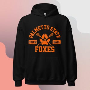 May include: Black hooded sweatshirt with an orange graphic design. The graphic features the text "Palmetto State" with a lacrosse stick and ball crossed over a paw print. The text "Stick" and "Ball" are below the sticks and the text "Foxes" is below the paw print.