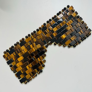 Op de afbeelding: Een decoratief oogmasker gemaakt van rechthoekige tijgeroogstenen en kleine bruine kralen. De stenen zijn gerangschikt in een baksteenpatroon, met bruine en zwarte tinten. Het masker is ontworpen om de ogen te bedekken.