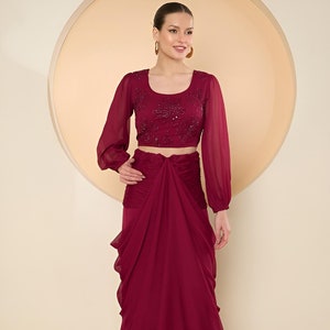 May include: A burgundy two-piece outfit. The crop top has long, sheer sleeves and sequin detailing on the bodice. The skirt is draped. The outfit is made of a flowing fabric. The outfit is suitable for special occasions.