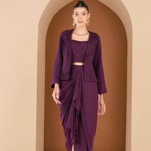 May include: A three-piece formal outfit in a deep purple hue. The ensemble includes a cropped top, a draped skirt with a high slit, and a matching jacket with beaded embellishments. The model is wearing gold earrings and a necklace.